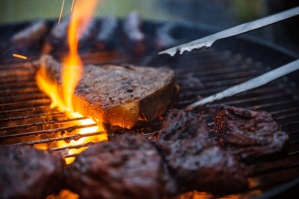 Flame grilled steaks being flipped on barbecue with tongs.